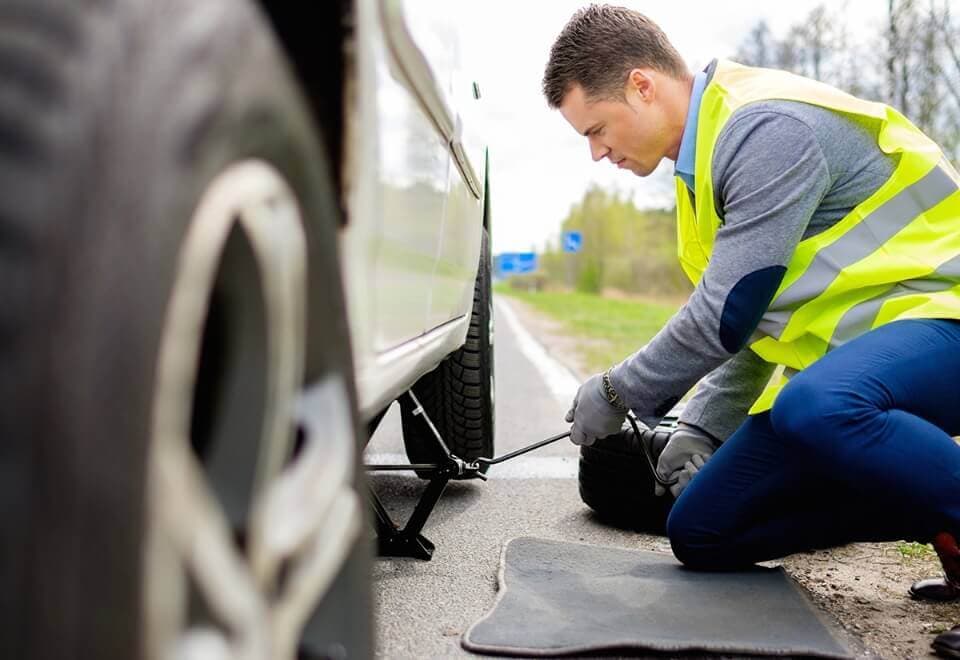 Picture this: A hardworking individual, clad in a bright reflective safety vest, crouches by the roadside, tirelessly changing a tire. They’re down on one knee, expertly wielding a tire iron to free those stubborn lug nuts while a jack securely hoists the vehicle just enough to get the job done. It's a scene as smooth and efficient as you'd expect from your trusted friends at DFW Towing Service—always ready to lend you a hand when you're in need!